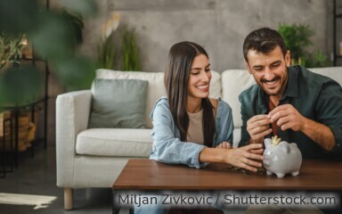 Eine Frau und ein Mann sitzen im Wohnzimmer vor dem Couchtisch, vor ihnen auf dem Tisch steht ein Sparschwein.
