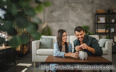 Eine Frau und ein Mann sitzen im Wohnzimmer vor dem Couchtisch, vor ihnen auf dem Tisch steht ein Sparschwein.