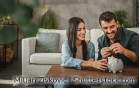 Eine Frau und ein Mann sitzen im Wohnzimmer vor dem Couchtisch, vor ihnen auf dem Tisch steht ein Sparschwein.